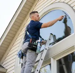 Professional window cleaner on a ladder using a squeegee to clean an exterior arched window, Charlotte, NC.